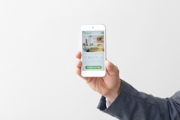 Businessman with mobile phone booking room in hotel online against light background