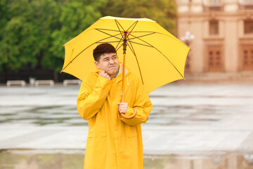 Young Asian man in raincoat and with umbrella on city street