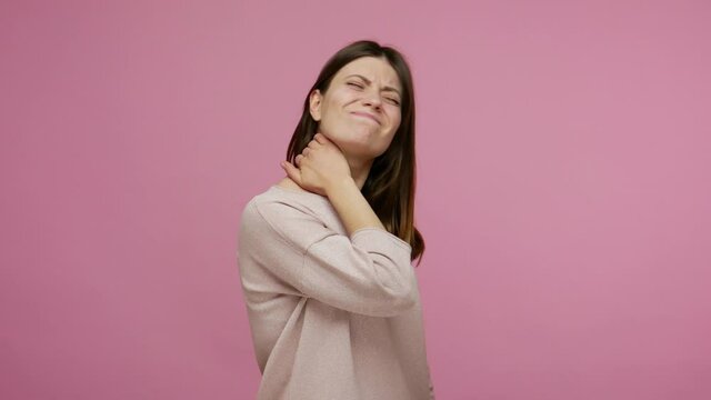 Sick tired young woman suffering intense pain in back neck, touching sore stiff muscles and frowning from ache, massaging shoulders feeling overworked. indoor studio shot isolated on pink background