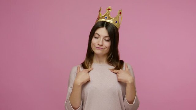 Look, I'm the boss! Arrogant selfish brunette woman wearing golden crown, symbol of power, looking with haughty proud expression, declaring her authority. studio shot isolated on pink background