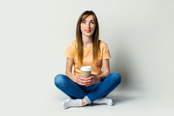 Young woman in casual wear holds a cup with coffee while sitting on the floor on a light background