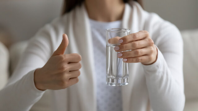 Close up image woman sit on couch holding glass of clean water showing thumbs finger up gesture, intake of enough quantity of aqua every day, improve metabolism, remove toxins, health benefits concept