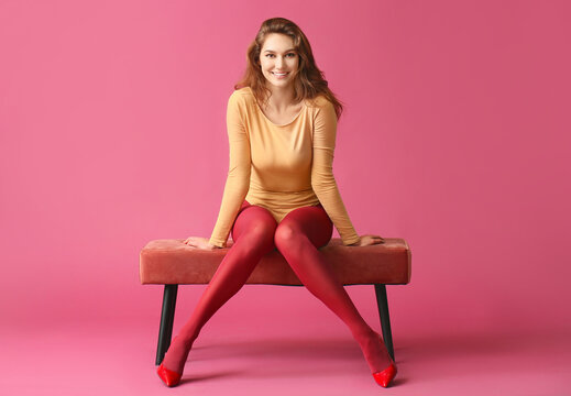 Beautiful young woman in tights sitting on bench against color background