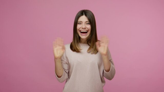 Lovely amiable brunette young woman waving hand to camera, greeting with hello gesture and smiling, welcoming saying hi with friendly expression. indoor studio shot isolated on pink background