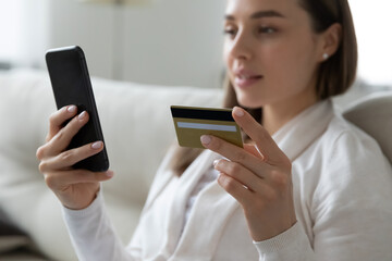 Close up focus female hands holding credit card and smartphone. Young woman sitting on sofa spend money at home, make order, buying using internet, commercial services, secure on-line payment concept