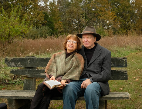 View of smiling senior couple sitting on bench in park on overcast day in fall, woman holds book on her lap
