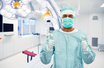 Fototapeta na wymiar medicine, surgery and people concept - indian male doctor or surgeon in mask and protective wear showing thumbs up over operating room at hospital background