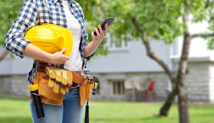 repair, construction and building concept - woman or builder with smartphone, helmet and working tools on belt over living house yard with garden background