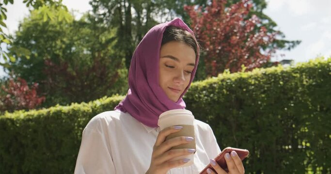 Beautiful young woman drink coffee and looking at smartphone, wearing traditional headscarf. Attractive Female in hijab