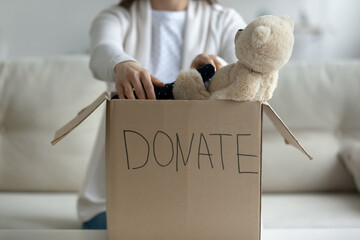 Woman sitting on couch at home put used clothes, soft toy in cardboard donation box close up image. Girl involved in aid nonprofit organization, selfless act of giving, generosity and charity concept