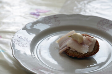 Slices of lard and a clove of garlic on a small hump of gray bread. Snack on a plate closeup.
