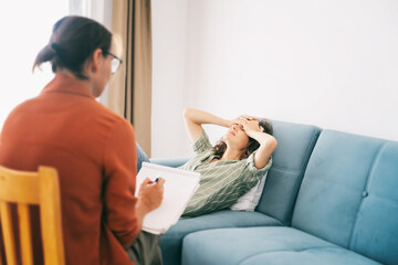 Depressed woman lying on sofa during psychotherapy session, psychologist making notes in her...