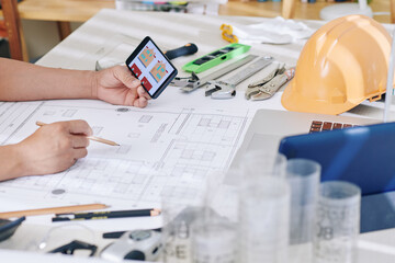 Construction engineer checking printed blueprint of building on screen of smartphone