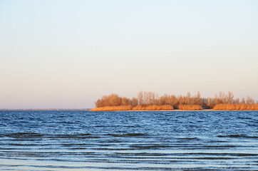Small waves on the bright blue surface of the water of the reservoir. A reddish island against a pale blue sky in the evening in early spring in March. Selective focus.