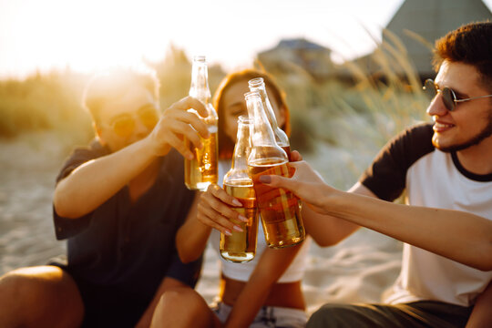 Young people sitting together at beach, drinking beer and having a party. Group of friends cheers with beers at the beach during summer vacation.