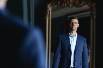 Handsome stylish man in blue jacket at home looking at mirror.