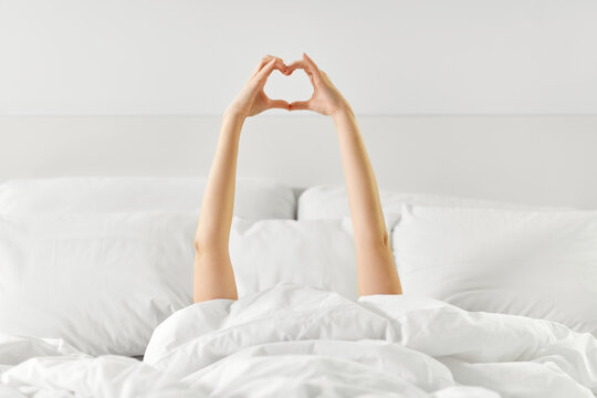gesture, comfort and morning concept - hands of young woman lying in bed making heart shape at bedroom