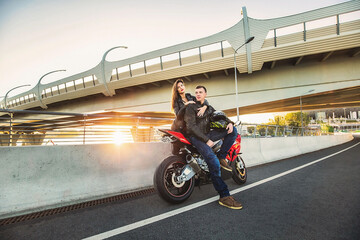 Young couple hugging while sitting on red sports motorcycle at bridge on sunset