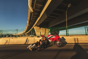 Young couple sitting on the asphalt road leaning on red sports motorcycle at bridge on sunset