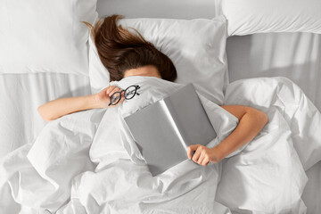 people, bedtime and rest concept - woman lying in bed under white blanket or duvet with book and...