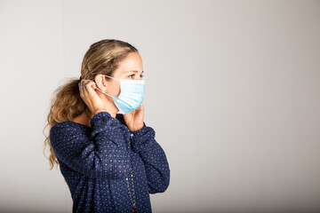 woman with blue shirt in medical mask adjusting the strap on the ear in front of white wall. 