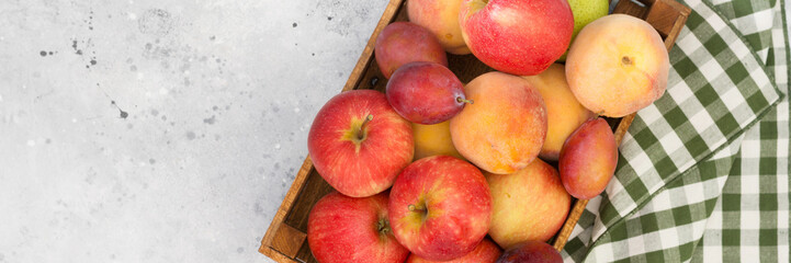 Various fruits in a wooden box on the light gray kitchen table. Apples, plums and peaches in a brown wooden box. Top view with space for text. Banner