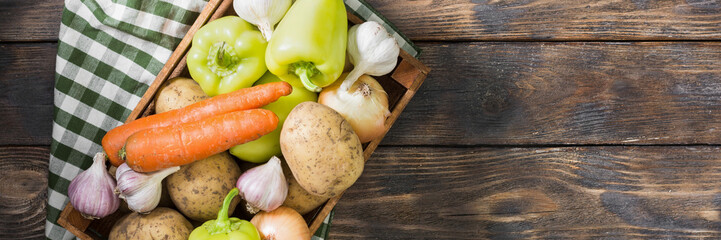 Various vegetables in a wooden box on a brown wooden table. Potatoes, peppers, onions, carrots and garlic in a brown wooden box. Top view with space for text. Rustic banner