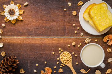Top view of soy milk in white cup with buttered bread on white plate and soy beans on wooden spoon Decorated with white flowers dried bark and pine cone with copy space on wooden background.