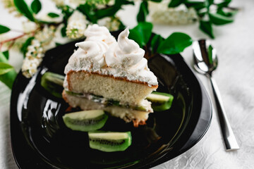 A slice of cake with meringue cream on a glossy black plate with slices of fresh kiwi. Cake decorated with a sprig of fragrant cherry blossoms