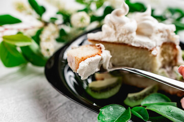 A slice of cake with meringue cream on a glossy black plate with slices of fresh kiwi. Cake decorated with a sprig of fragrant cherry blossoms