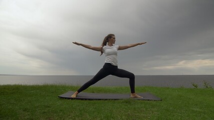 Fototapeta na wymiar Young fit woman practice yoga on coast near the lake or sea. Woman doing Warrior II Pose