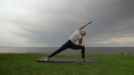 Fototapeta na wymiar Young fit woman practice yoga on coast near the lake or sea. Woman doing Revolved Warrior with Arms Extended Pose