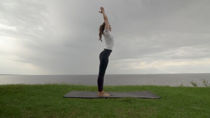 Young fit woman practice yoga on coast near the lake or sea. Woman doing Mountain Arms Up Pose