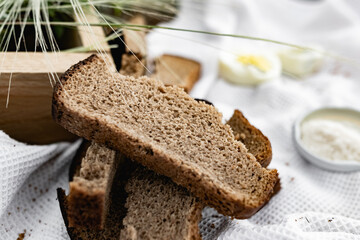 Slices of black bread and a boiled chicken egg on a white towel on the green grass. Picnic in nature with simple food. Next to the salt and flowers with plants in a wooden box.