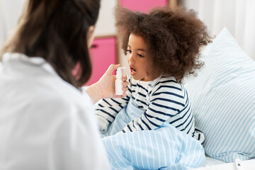 healthcare, medicine and people concept - doctor with oral spray treats little sick african american girl in bed at home