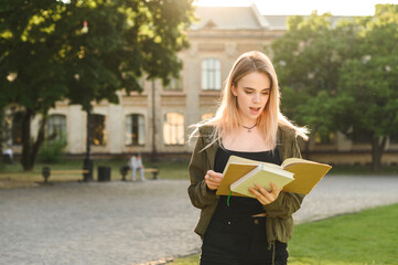 Surprised attractive student girl standing in university campus park, reading a book with surprised face. Shocked girl looks at exercise book and learns to break at school.