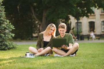 Beautiful couple of students sitting on grass in university campus and using laptop with smile on...