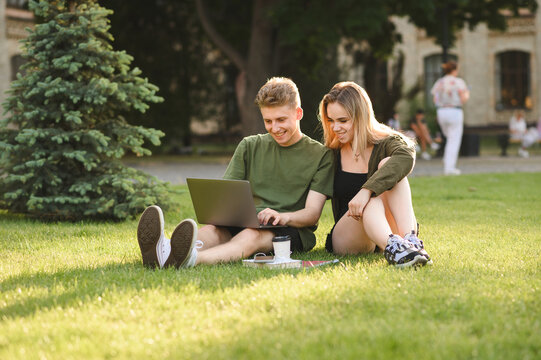 Couple of happy students using laptop while sitting on university campus lawn, looking at screen and smiling. Smiling girl and young man studying with laptop at park break. Student leisure.