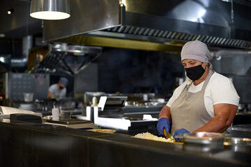 cooking, profession and people concept - woman in age chef wearing protective black mask on her face while working in restaurant kitchen