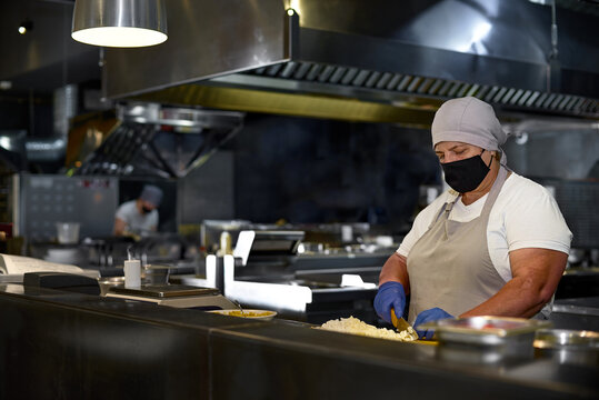 Cooking, Profession And People Concept - Woman In Age Chef Wearing Protective Black Mask On Her Face While Working In Restaurant Kitchen