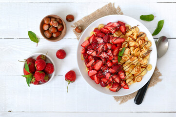 Pancake cereal in a bowl with fresh strawberries and nuts on a white wooden background. Tasty and trendy breakfast. Organic Dutch Mini-Pancakes. The top view