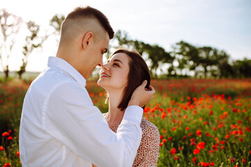 Romantic young couple hug among red beautiful poppy field. Loving boy and girl stand, hug and kiss showing their feelings to each other. Freedom, wedding, family and love concept.