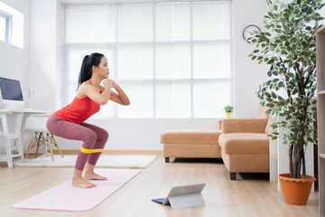 Obraz premium Asian woman exercising at home on a yoga mat. She uses rubber resistance. She's doing squats.