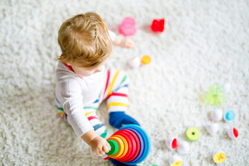 Fototapeta na wymiar Adorable cute beautiful little baby girl playing with educational toys at home or nursery. Happy healthy child having fun with colorful wooden rainboy toy pyramid. Kid learning different skills.