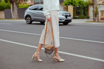Detail of young fashionable woman wearing white sweatshirt, pants and gold high heels. She holding stylish beige handbag with silver details in hands. Street style.