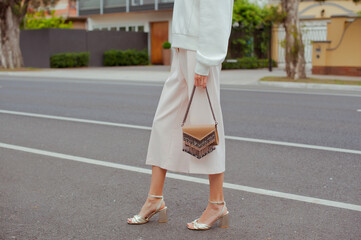 Detail of young fashionable woman wearing white sweatshirt, pants and gold high heels. She holding stylish beige handbag with silver details in hands. Street style.