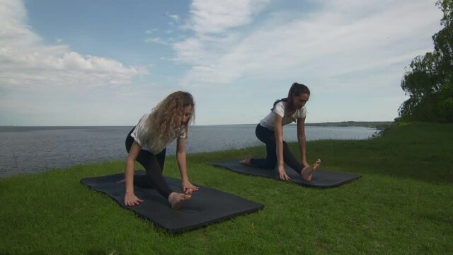 Two young women practice yoga on coast near the lake or sea. Training Crescent Lunge on the Knee pose.