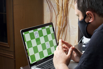A man in a medical mask solves a chess problem online. Chess board with figures on a laptop screen. Leisure in self-isolation. International chess day.