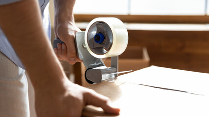 Naklejka premium Close up man using adhesive tape dispenser, packing cardboard box with belongings or parcel, moving day and relocating delivery service concept, young male preparing to relocation
