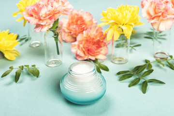 Face moisturizer cream and pink rose carnations and yellow chrysanthemums or mums flowers on pastel canvas blue background. Flat lay or top view, copy space for your text